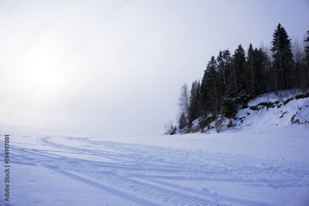 winter road (frozen river with a highly wooded shore) with traces of snowmobiles, with a horizon, lost in a frosty haze