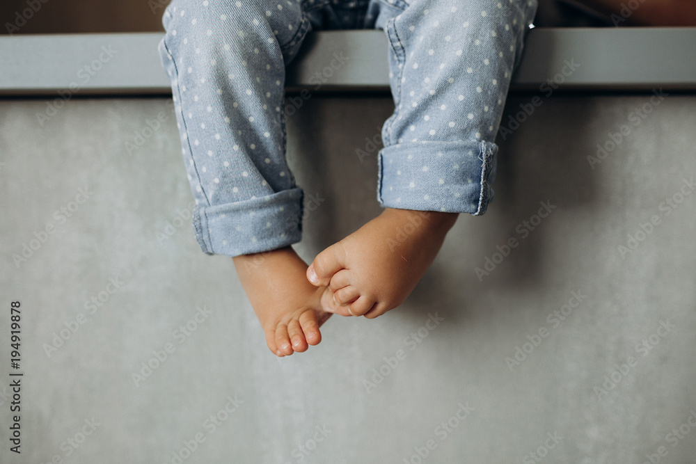 close-up of a baby's legs in blue jeans that hang over a gray wall background