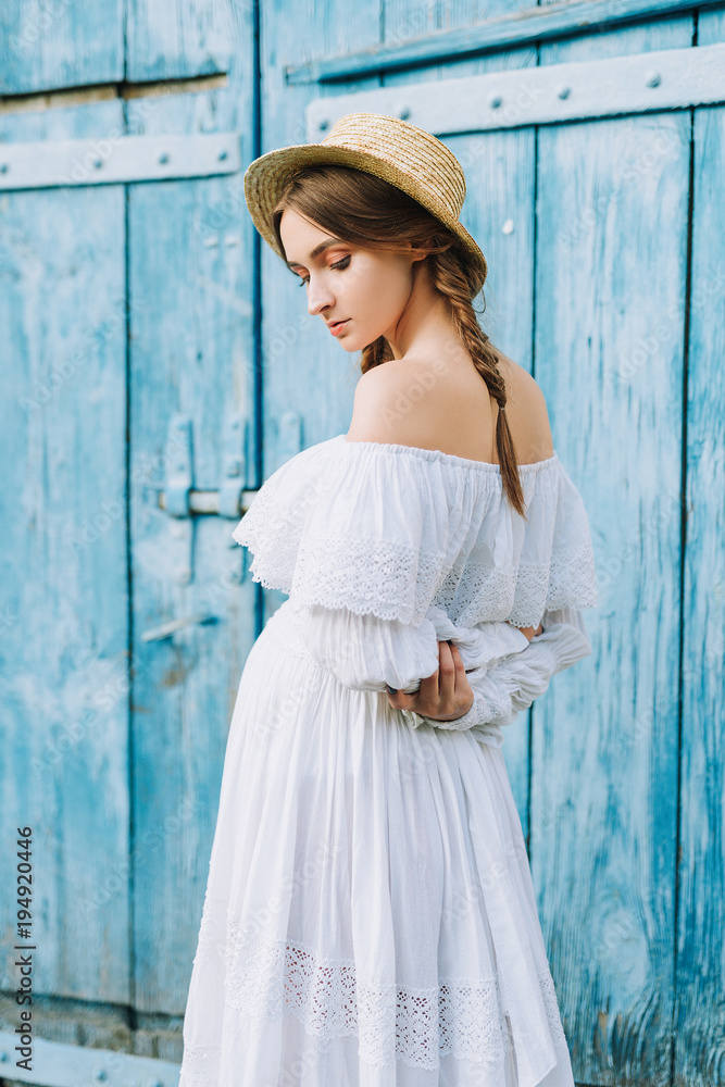 Fototapeta premium portrait of a pregnant girl in a white long dress and a straw hat that behind her back has brought her hands against the blue door