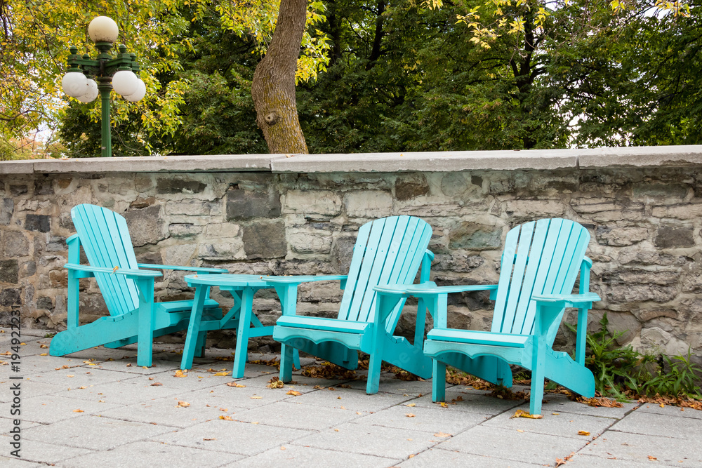 Three wooden turquoise Adirondack chairs with a small wooden table in front of a stone wall with trees and a lamp post.