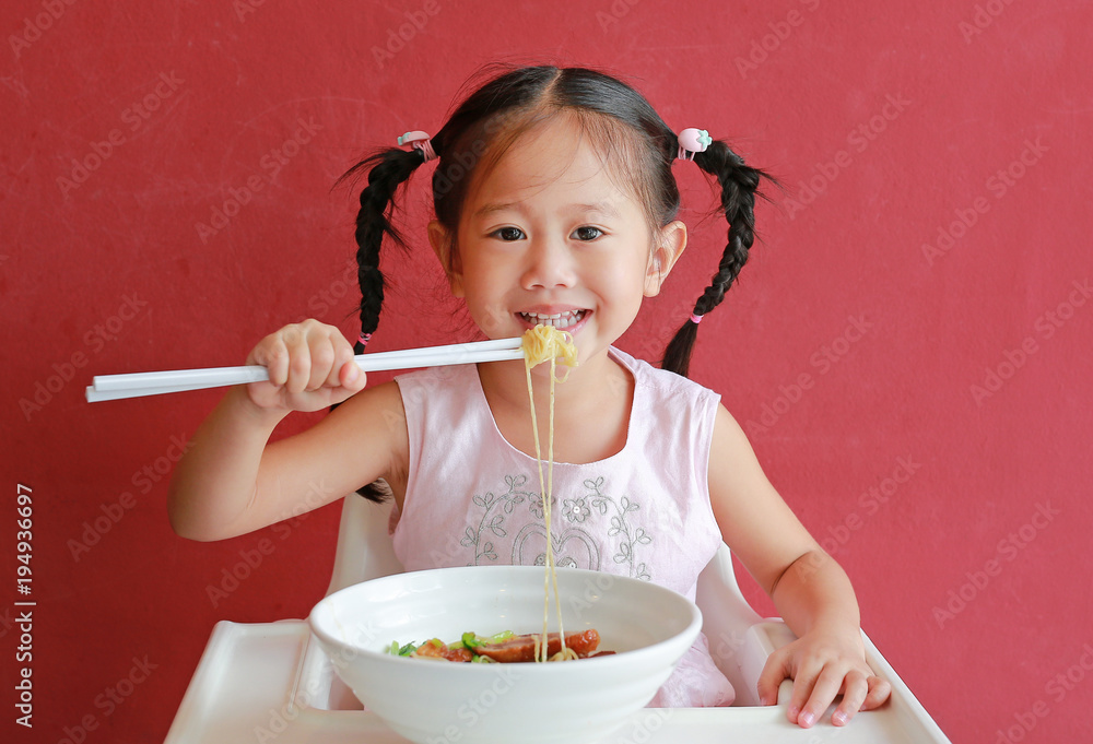Happy little asian girl eating Egg noodle on high chair against red wall background. Happy little asian girl eating Egg noodle on high chair against red wall background.