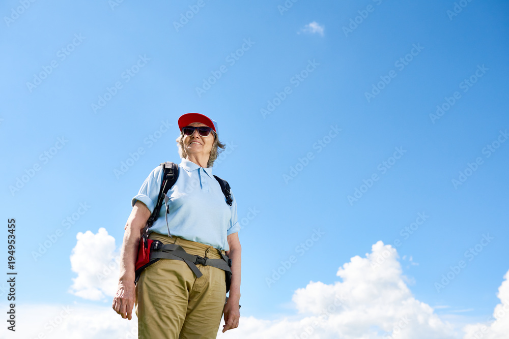 Low angle portrait of active senior woman travelling on hiking trip, posing smiling happily against clear blue sky, copy space Low angle portrait of active senior woman travelling on hiking trip, posing smiling happily against clear blue sky, copy space