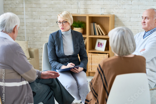 Portrait of blonde female psychiatrist wearing glasses leading group therapy session for senior people in retirement home, copy space