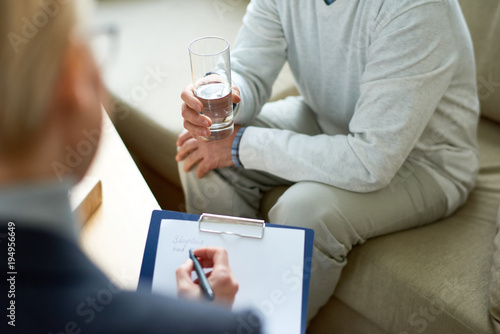 High angle close-up of female psychologist writing on clipboard while listening to depressed senior patient with glass of water