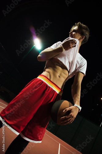 Sweaty and confidant. Handsome young man in sportswear holding basketball and pulling his t-shirt while standing on basketball court  © MARIIA