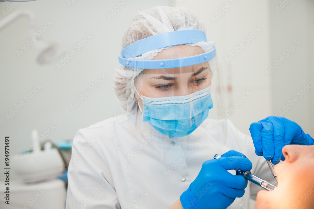 Female stomatologist in blue protective mask examining patient's teeth. Dentist caries treatment at dental clinic office. People, medicine, stomatology and health care concept Female stomatologist in blue protective mask examining patient's teeth. Dentist caries treatment at dental clinic office. People, medicine, stomatology and health care concept
