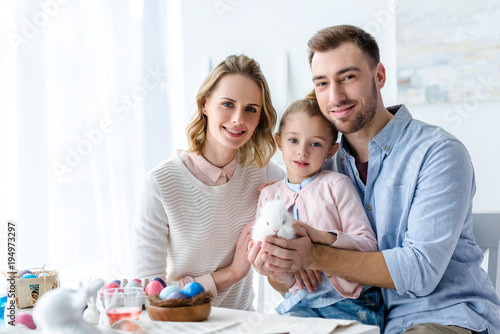 Happy family holding Easter rabbit with painted eggs on table