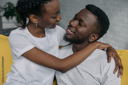 young african american couple in white t-shirts hugging and smiling each other at home