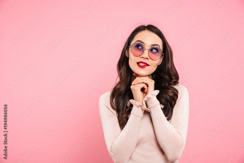 Photo of dreaming adult girl with red lips wearing trendy sunglasses looking upward keeping hands together, isolated over pink background Photo of dreaming adult girl with red lips wearing trendy sunglasses looking upward keeping hands together, isolated over pink background