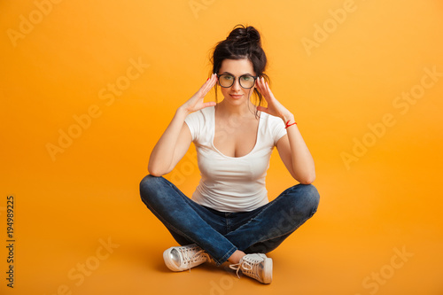 Young woman 20s in casual clothing and eyeglasses posing on camera with adorable look while sitting with legs crossed on the floor, over yellow wall