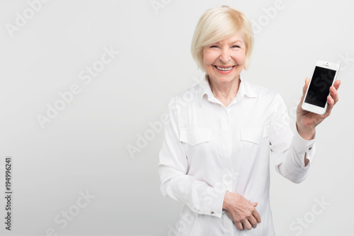 Smiley senior woman is holding a new smartphone in her hand. She has learned how to use it. She is passioned about internet and new technologies. Isolated on white background.