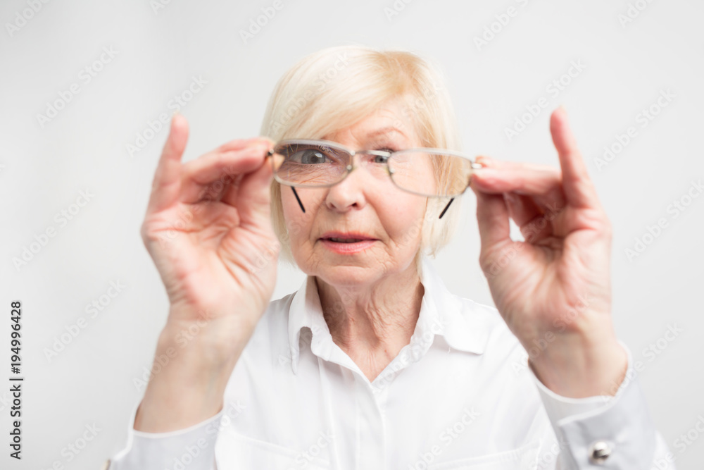 Close up of old woman is very attentive to details. She is looking to her glasses trying to find dirty spots there. She likes all things to stay clean. Isolated on white background.