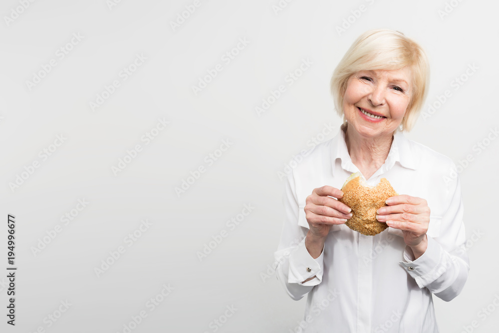 Old but satisfied woman is holding a burger in her hands. She has just made one bite. This lady likes the taste of this meal. Sometimes she likes to eat some junk food. Isolated on white background. Old but satisfied woman is holding a burger in her hands. She has just made one bite. This lady likes the taste of this meal. Sometimes she likes to eat some junk food. Isolated on white background.