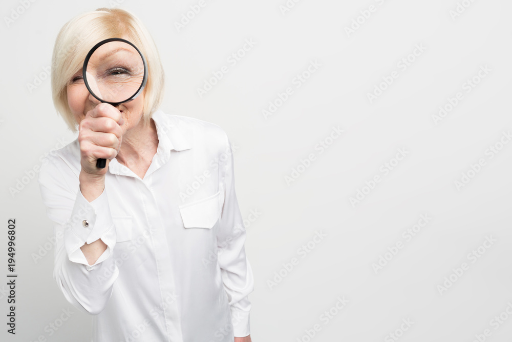 Close up of suspicious old woman in white blouse looking straight ahead through the reading glass. She looks strange. Cut view. Isolated on white background. Close up of suspicious old woman in white blouse looking straight ahead through the reading glass. She looks strange. Cut view. Isolated on white background.