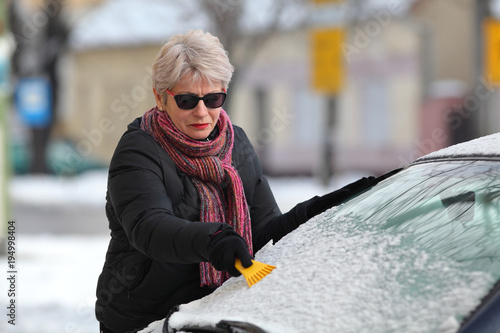 Winter scene, female driver cleaning snow from  windshield of car using scraper 