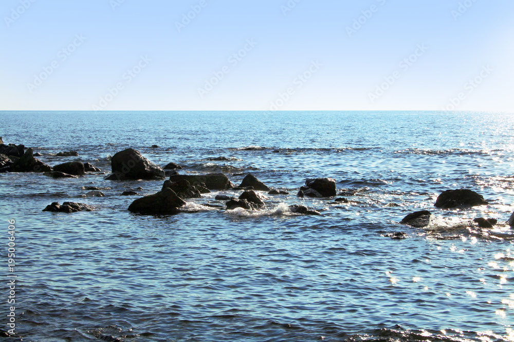 Obraz premium Big stones in the sea. Big stones not far from the beach form breakwaters. Small waves reflect the blue sky in different nuances.