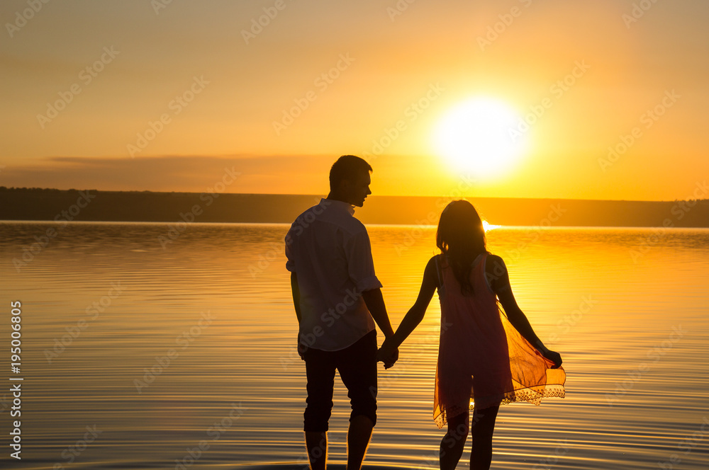 Young couple is walking in the water on summer beach. Sunset over the sea.Two silhouettes against the sun. Just married couple hugging. Romantic love story. Man and woman in holiday honeymoon trip.