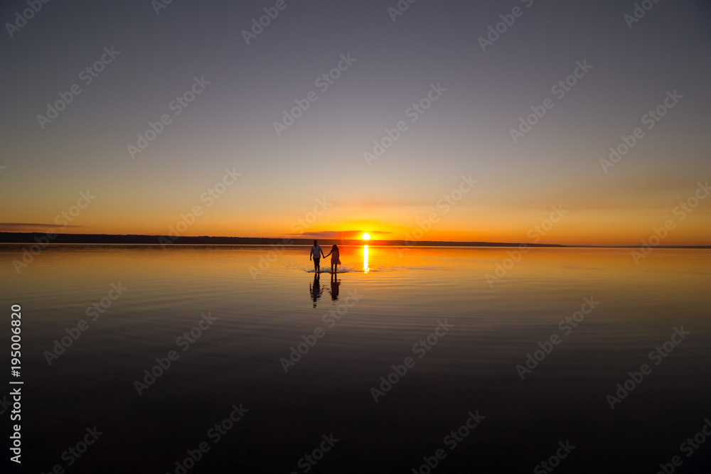 Young couple is walking away in the water on summer beach. Sunset over the sea.Two silhouettes against the sun. Just married couple in romantic love story. Man and woman in holiday honeymoon trip.