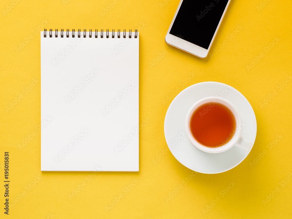 Top view of modern bright yellow office desktop with blank notepad, cup of tea, smartphone. Mock up, empty space Top view of modern bright yellow office desktop with blank notepad, cup of tea, smartphone. Mock up, empty space