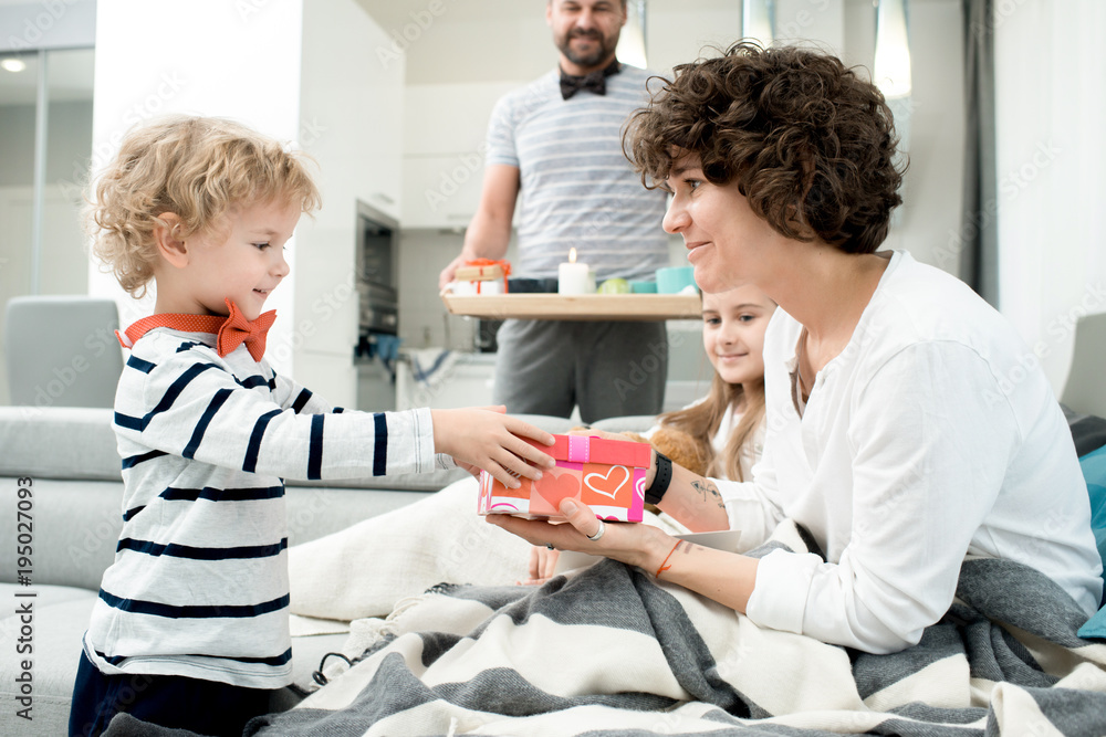 Obraz premium Side view portrait of adorable little boy giving present to his mom on Valentines day, with the rest of happy family in background
