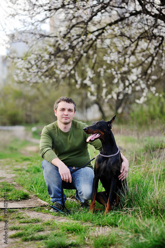 Young man sits on the grass with dobermann dog on a background of spring blooming trees