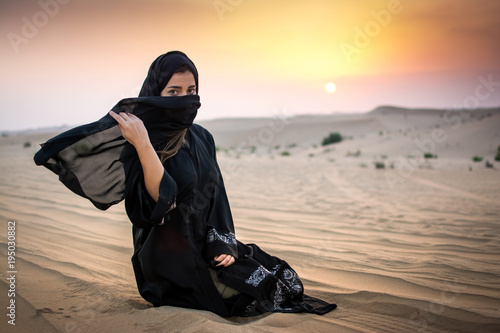 Portrait of Muslim woman sitting on sand in the desert during sunset. Beautiful young woman holding veil to cover her face.