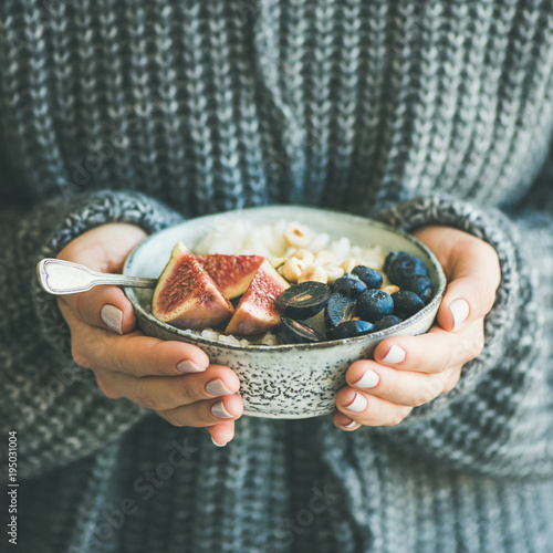 Healthy winter breakfast. Woman in woolen sweater holding bowl of rice coconut porridge with figs, berries, hazelnuts, square crop. Clean eating, vegetarian, vegan, alkiline diet food concept