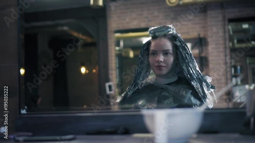 Beautiful woman with dye on her hair is sitting in hair salon, turning on chair, looking in the miroor and smiling to herself. Slow mo photo