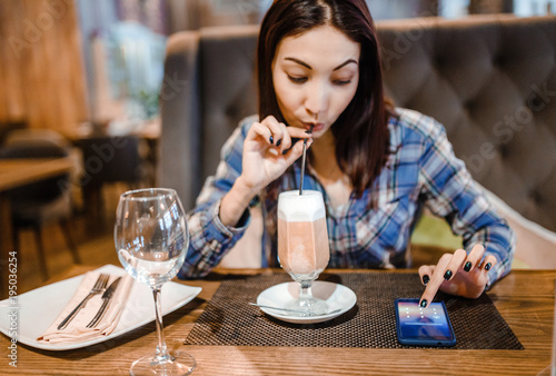 woman sitting at a wooden table in a cafe  drinking coffee and holding a smartphone.