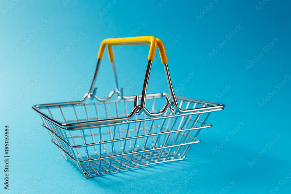 Close up of metal grocery basket for shopping in supermarket with raised handles and yellow plastic elements isolated on blue background. Concept of shopping. Copy space for advertisement Close up of metal grocery basket for shopping in supermarket with raised handles and yellow plastic elements isolated on blue background. Concept of shopping. Copy space for advertisement