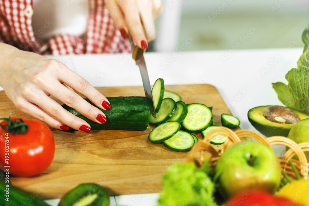 Caucasian girl with neat red manicure cutting cucumber on chopping board, wearing checked red shirt and white t-shirt, sitting at the table full of green and healthy food, closeup Caucasian girl with neat red manicure cutting cucumber on chopping board, wearing checked red shirt and white t-shirt, sitting at the table full of green and healthy food, closeup