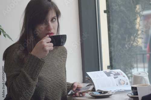 beautiful girl drinking tea and using a mobile phone while sitting in a cafe in front of a large window