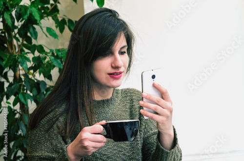 beautiful girl drinking tea and using a mobile phone while sitting in a cafe in front of a large window