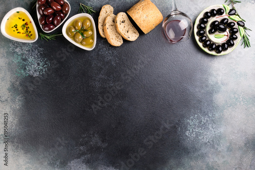 Olives, red wine, ciabatta bread, oil, herbs and spices on black stone background. Mediterranean snacks. Appetizer gourman food. Copy space, top view