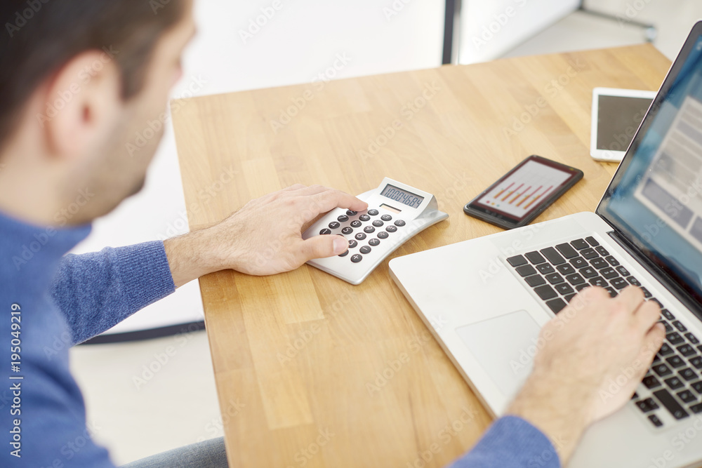 Managing his small business. Rear view shot of businessman counting on calculator and typing on laptop's keyboard. Managing his small business. Rear view shot of businessman counting on calculator and typing on laptop's keyboard.
