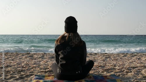 Girl in warm clothes sits on the shore of the Mediterranean Sea on a cool autumn day.
