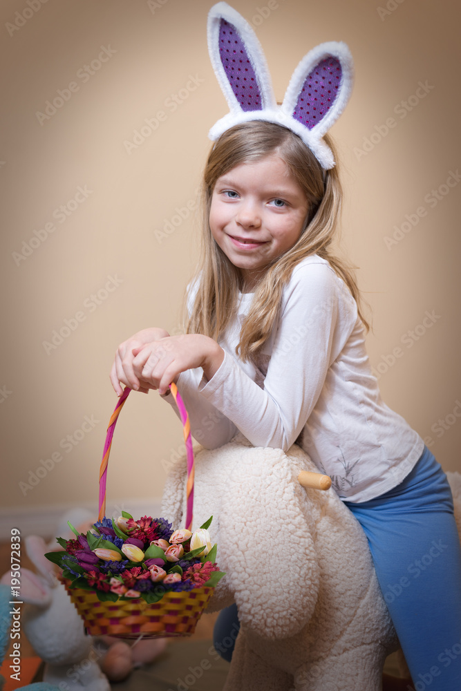 Cute little Caucasian blonde girl in Easter Bunny costume  is sitting on fluffy bunny toy and keeping basket with  flower