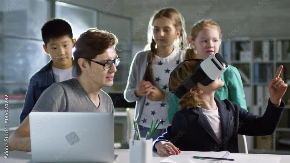 Group of four primary school children, one of them wearing VR glasses, learning to use virtual reality with their teacher Group of four primary school children, one of them wearing VR glasses, learning to use virtual reality with their teacher