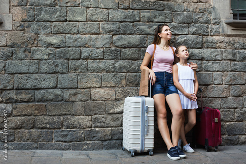 Woman and little girl traveler with suitcase leaning against stone wall