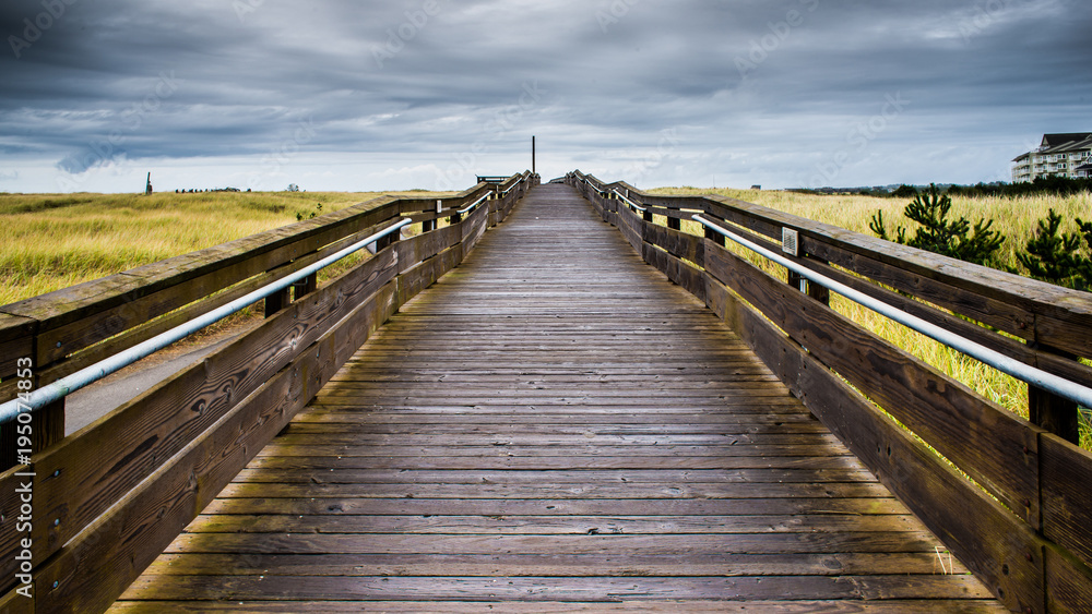 A Wood Bridge Leads To The Horizon By The Horizon Above Large Green Tall Grass Field Under The Stormy Sky And Rolling Clouds In Long Beach Washington
