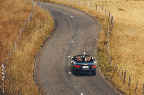 Top view. Happy just married couple is driving a convertible car on a country road for their honeymoon, the bride have fun with hands up