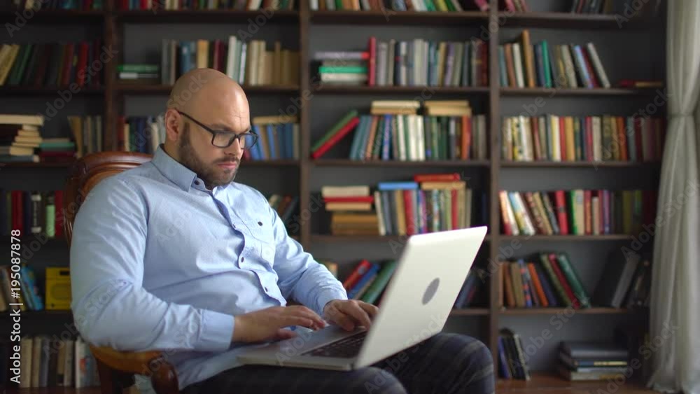 the scientist works at home laptop on the background of the library. businessman or scientist or architect using laptop. Thoughtful male scientist typing new article on laptop, freelance work. 4 k the the scientist works at home laptop on the background of the library. businessman or scientist or architect using laptop. Thoughtful male scientist typing new article on laptop, freelance work. 4 k the