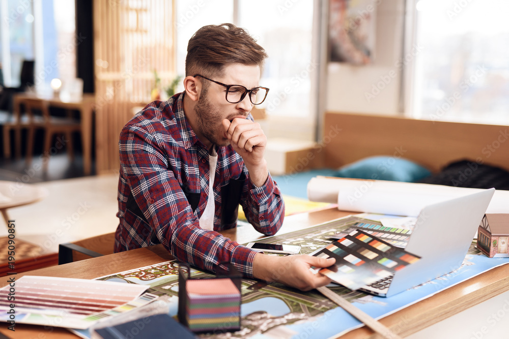 Freelancer man looking at color swatches at laptop sitting at desk.