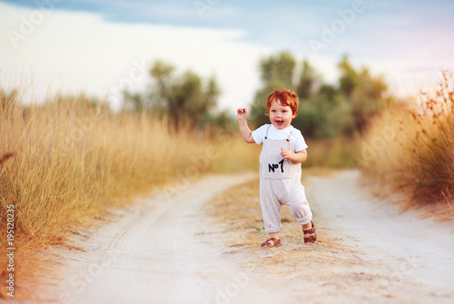 adorable redhead toddler baby boy in jumpsuit running along rural summer road in sunburned field