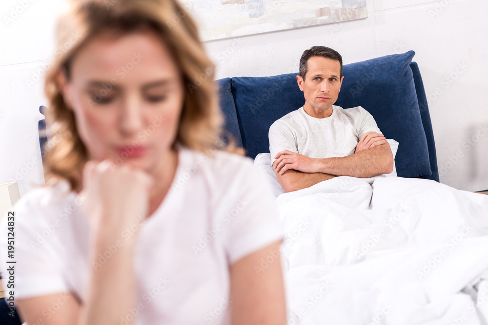 Fototapeta premium selective focus of upset woman sitting on bed with husband behind at home, relationship difficulties concept
