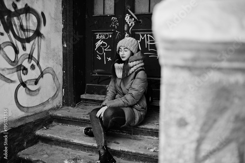 Portrait of brunette girl in gray scarf and hat, glasses sitting against urban entrance.