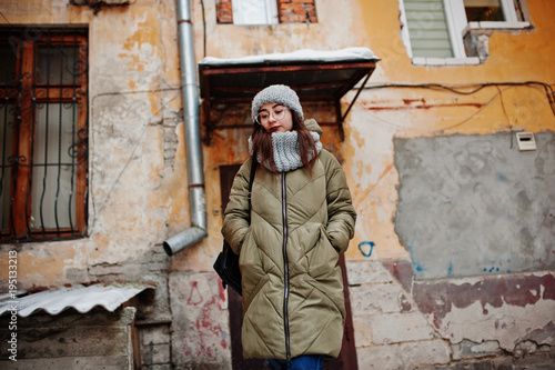 Portrait of brunette girl in gray scarf and hat, glasses at cold weather against orange wall of old house.