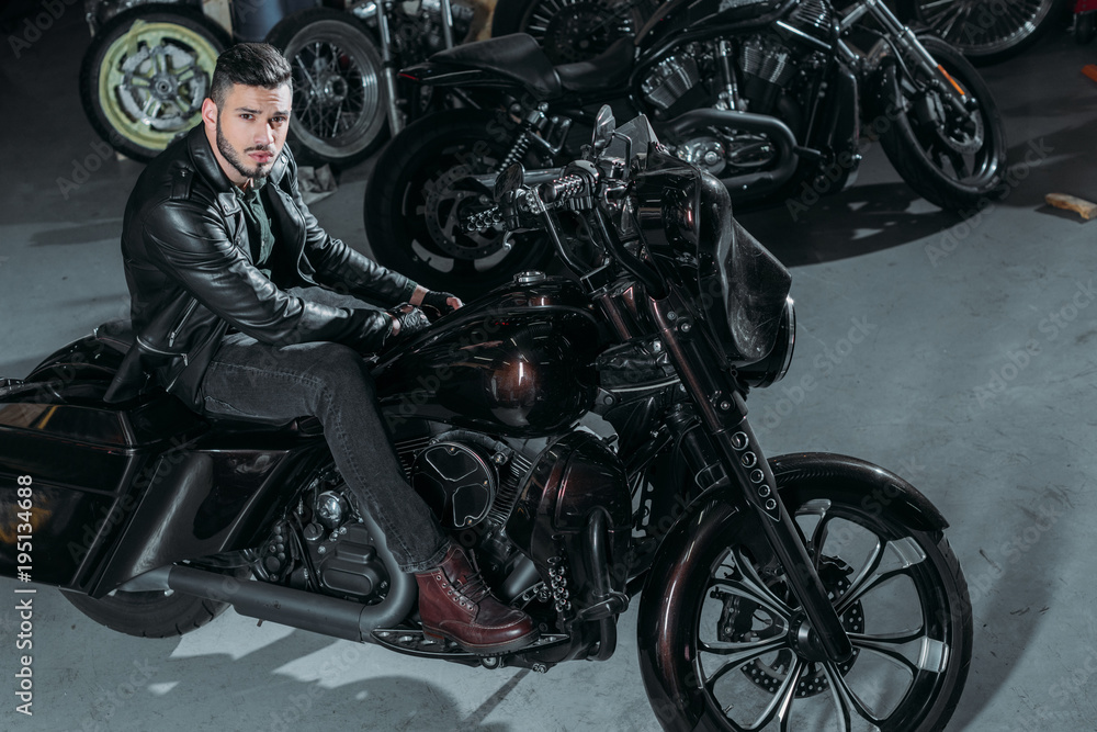 high angle view of handsome young man in leather jacket sitting on bike at garage high angle view of handsome young man in leather jacket sitting on bike at garage