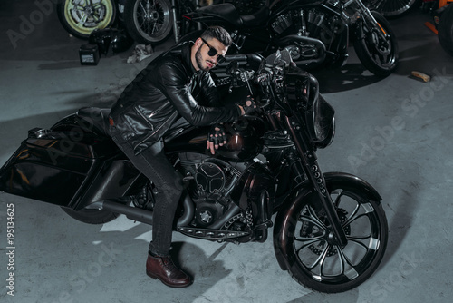 high angle view of handsome young man in sunglasses and leather jacket on bike at garage