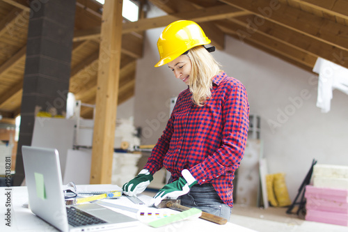 young, pretty, blonde woman with yellow helmet and green gloves is planning on the notebook and tablet the expansion of her loft